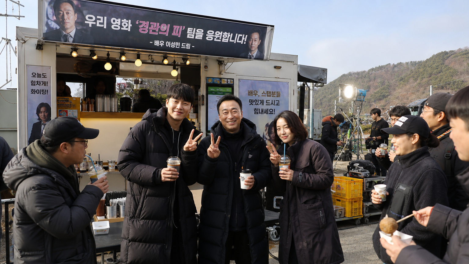 야외에서 다채로운 배너 아래서 한국 배우와 스태프들이 다과를 즐기는 활기찬 촬영장의 모습