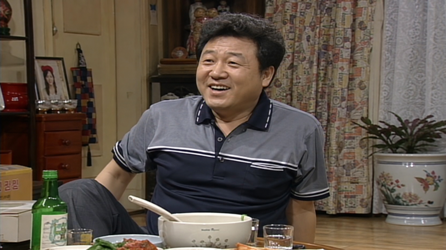 활발하고 유머 넘치는 분위기의 한국 시트콤 장면 속 코믹한 아빠 역할