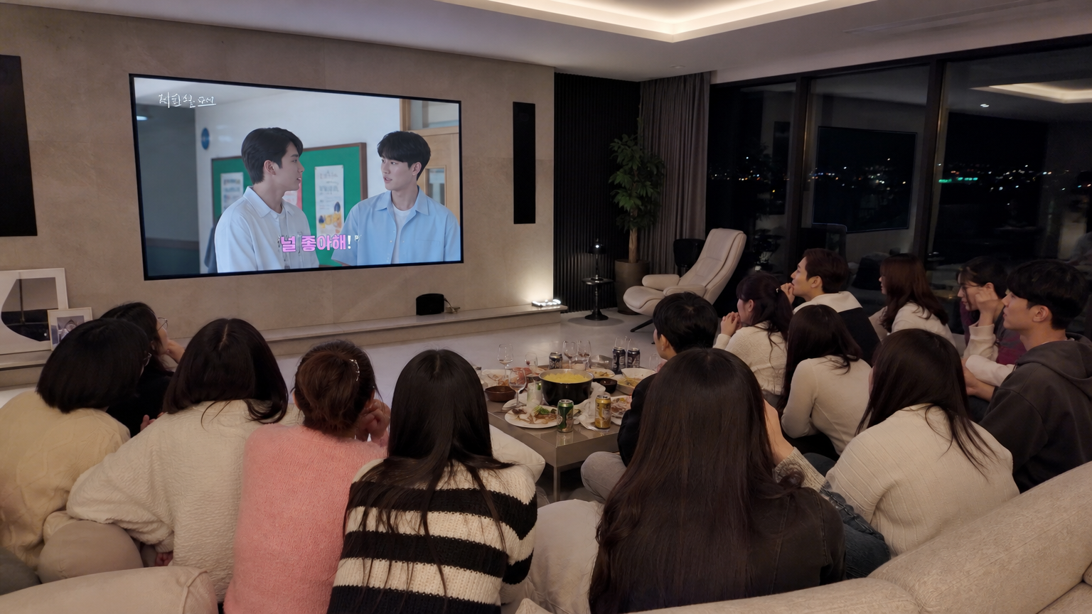 현대적인 거실에서 대형 화면으로 한국 리얼리티 쇼를 보는 많은 시청자들의 모습