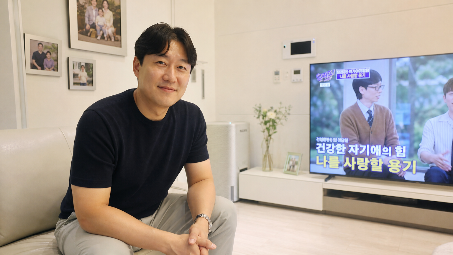 캐주얼한 복장을 입고 가족사진이 걸린 거실에 앉아 있는 중년 남성