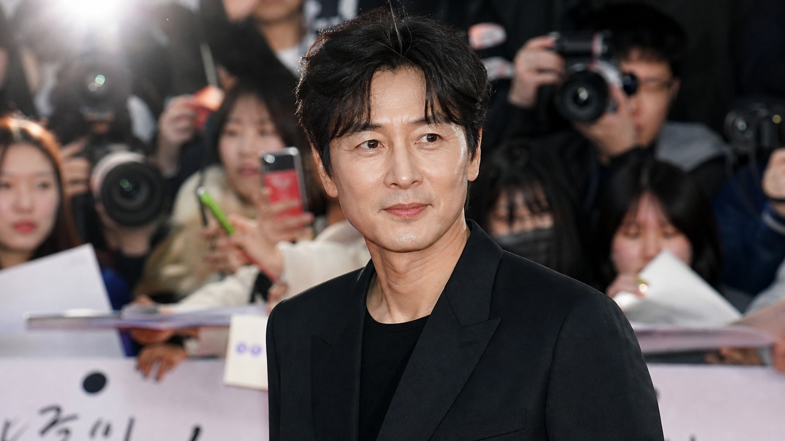 중년의 배우가 행사장에서 젊고 자신감 넘치는 모습으로 대중 앞에 서 있는 장면