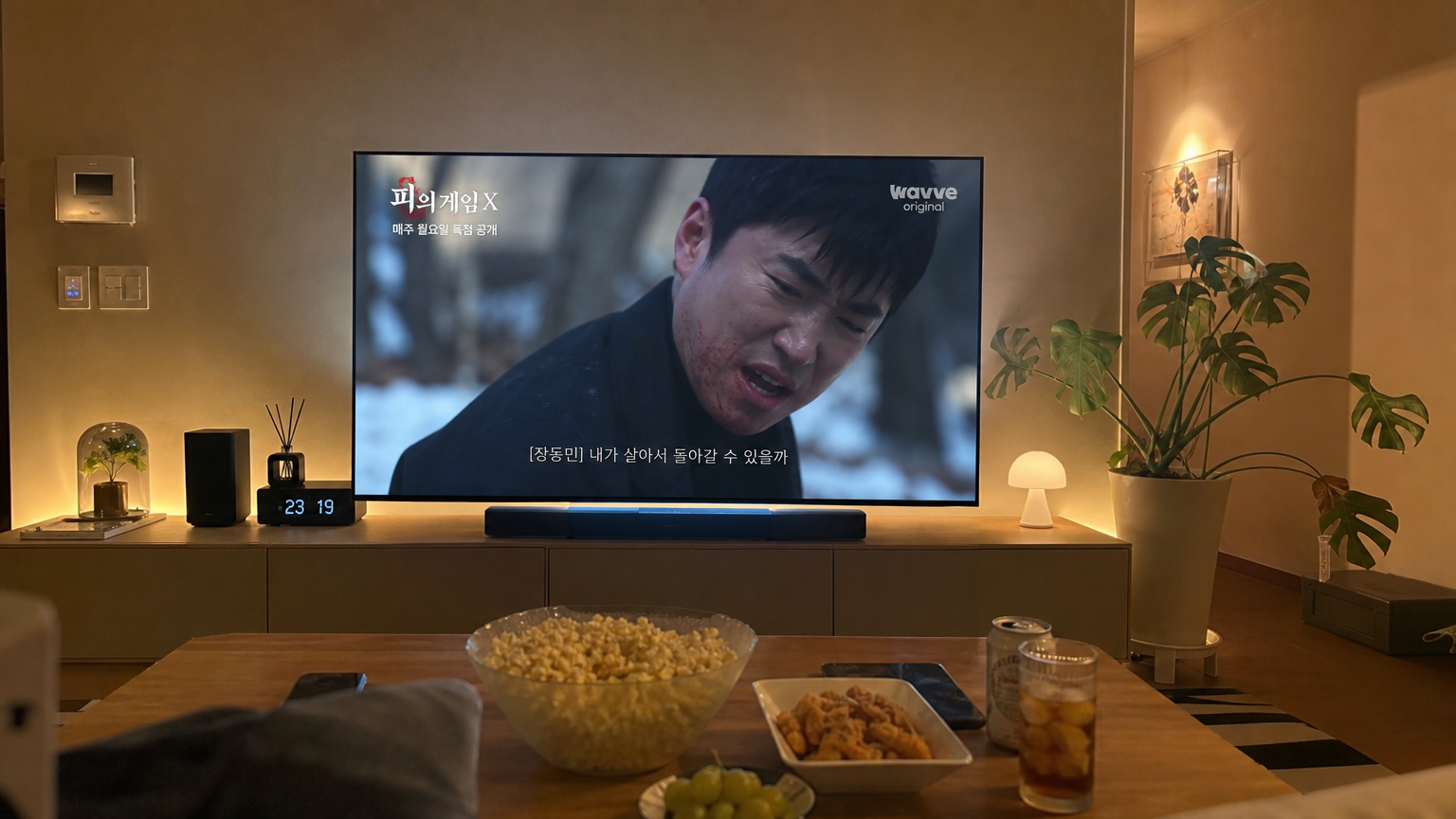 현대적인 거실에서 대형 스크린에 비친 '피의게임X' 장면과 함께 간식들이 놓인 아늑한 분위기