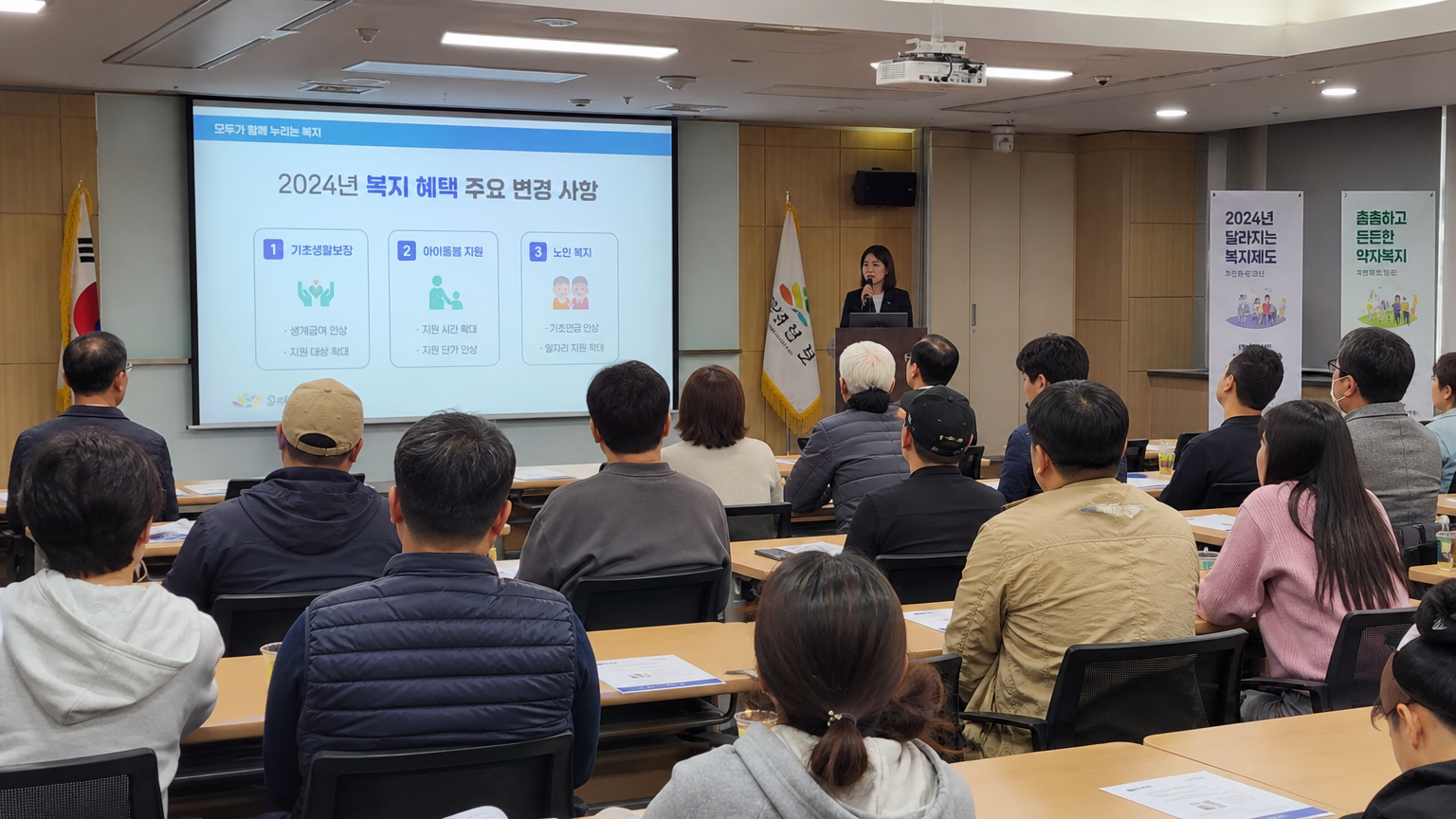 다양한 한국인들이 회의실에서 2024년 복지 제도 변화 발표를 듣고 있는 장면