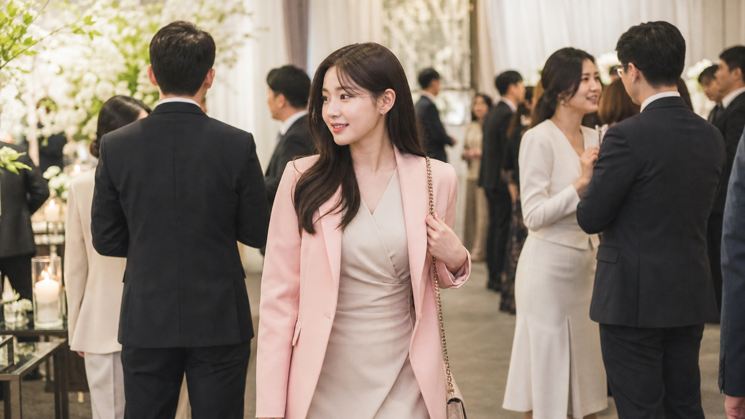 베이지색 드레스와 파스텔 핑크 재킷을 입고 있는 여성이 결혼식장에서 다른 하객들과 어우러져 있는 장면