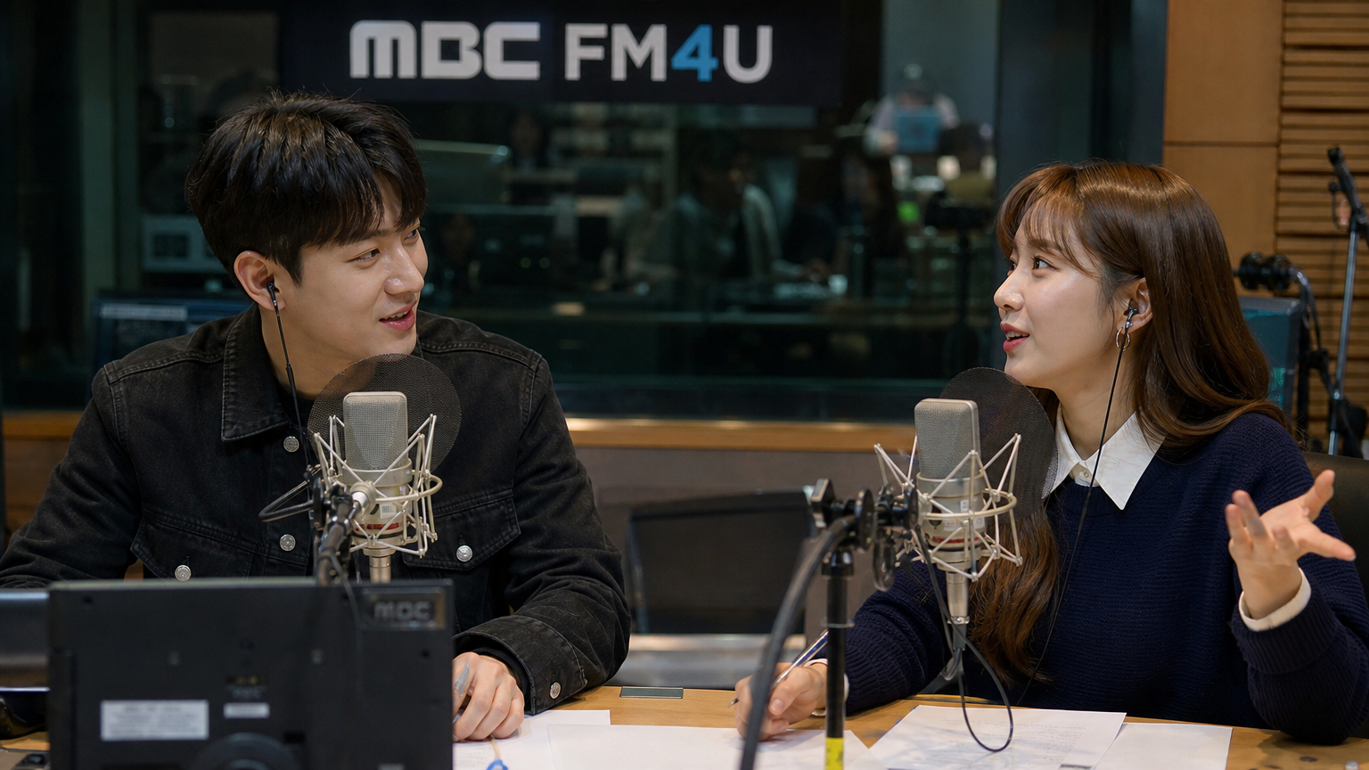 라디오 스튜디오에서 마이크를 앞에 두고 활발하게 대화하는 두 명의 한국 라디오 진행자