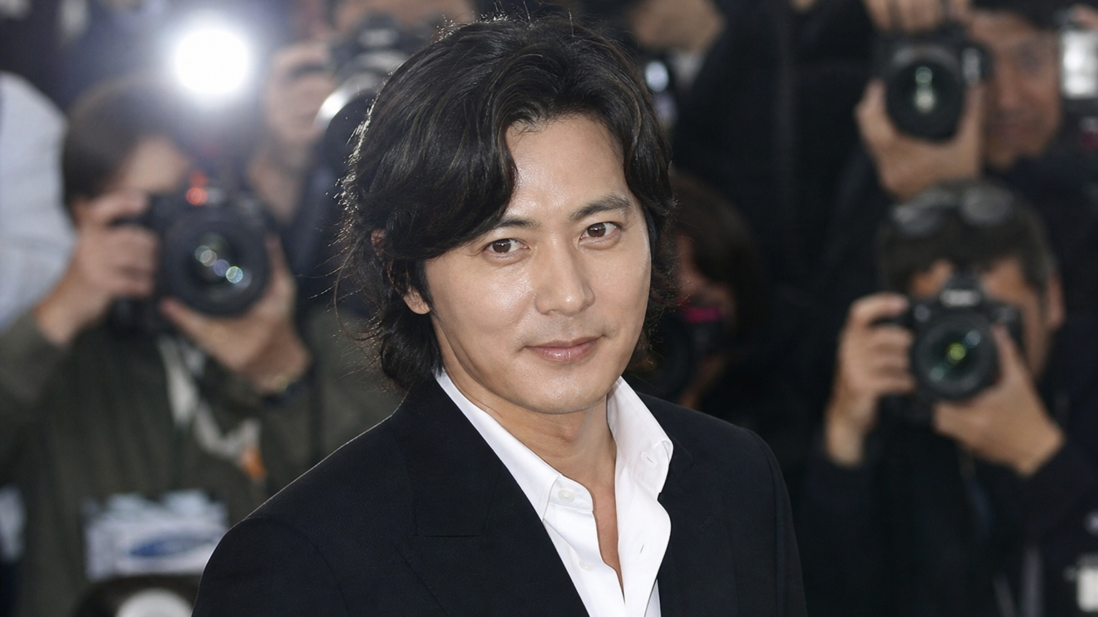 배우 장동건이 공식 행사에서 젊고 스타일리시한 모습으로 카메라 플래시를 받는 장면