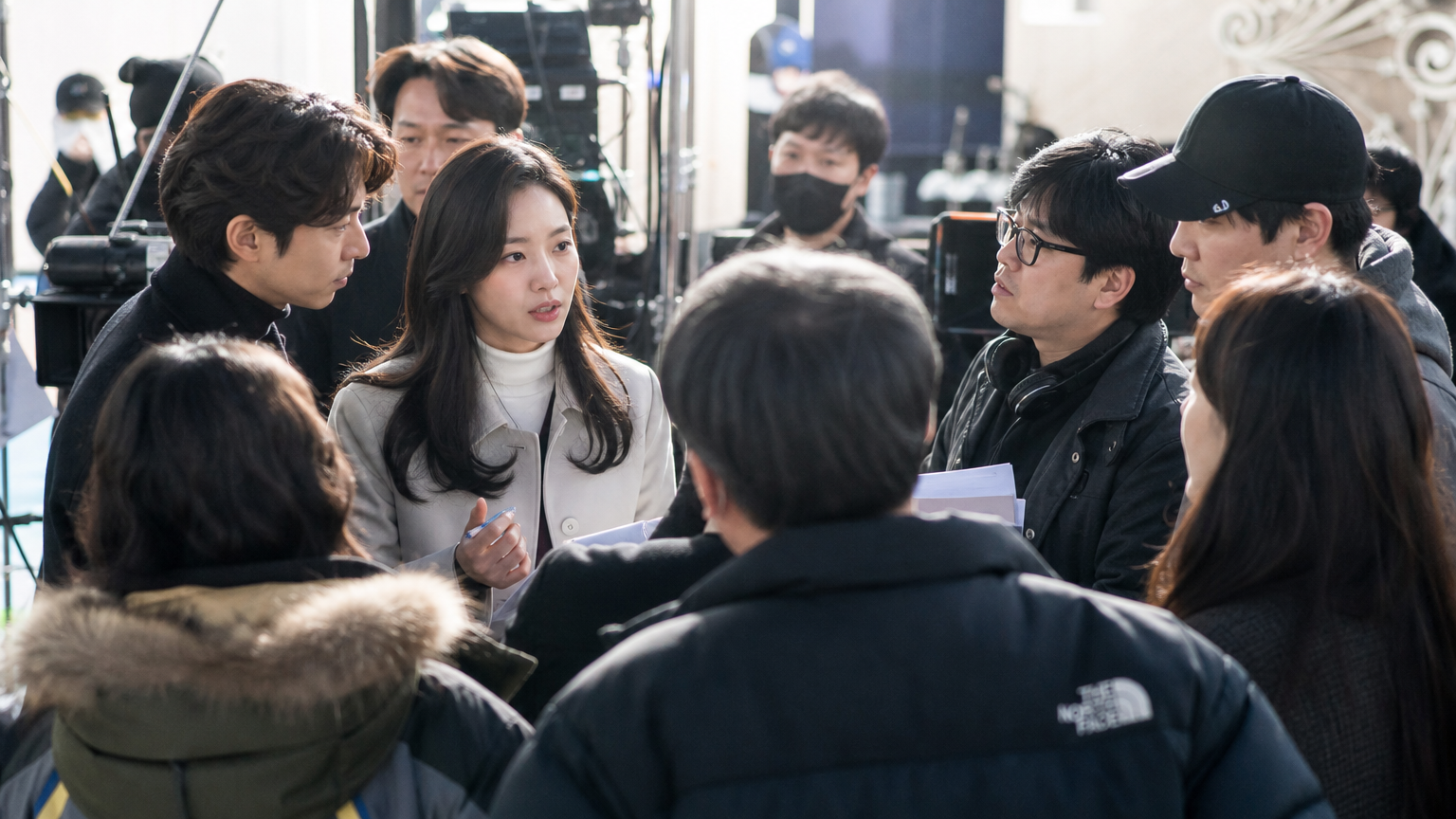 한국 드라마 세트장에서 배우들이 제작진과 활발히 대화하는 장면