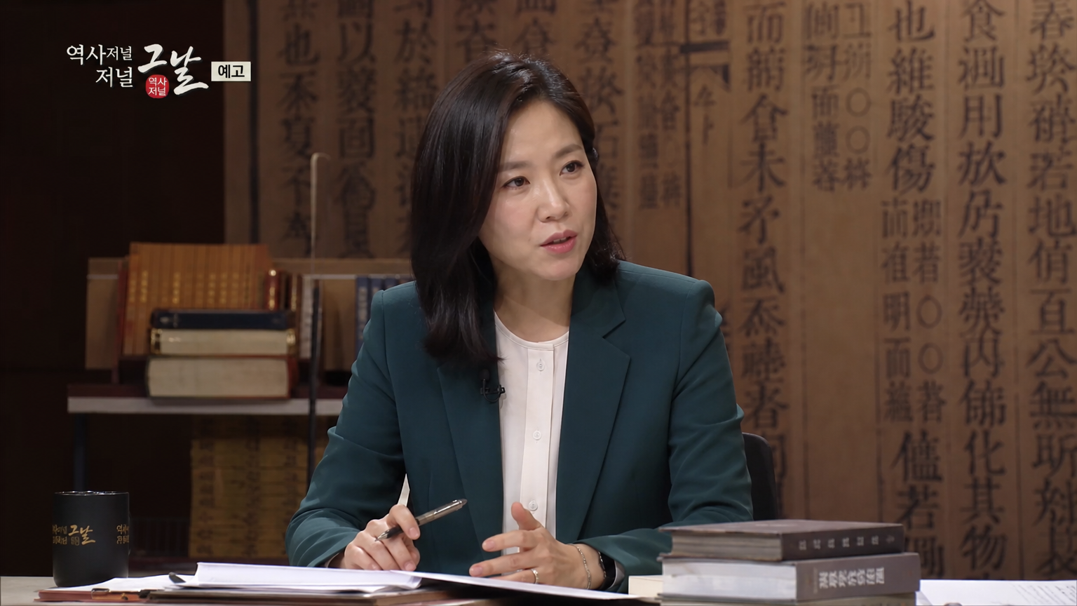 역사 토론 프로그램에서 책과 스크립트 사이에 앉아 있는 지적인 표정의 한국 여성 패널리스트
